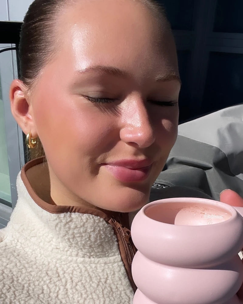 A person with closed eyes smiles while holding a pink mug. They are wearing a light-colored fleece jacket and gold hoop earrings. Sunlight illuminates their face, and a blurred background suggests an outdoor setting.